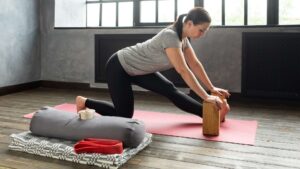 Akcesoria do jogi: wałek, klocek, pasek, koc do jogi,. Zdjęcie przedstawia kobietę podpierającą się o klocek do jogi