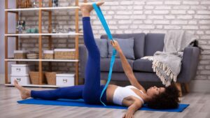 Zdjęcie przedstawia kobietę ćwiczącą jogę. Leży na plecach na macie do jogi i podnosi nogę podpierając ją paskiem do jogi
